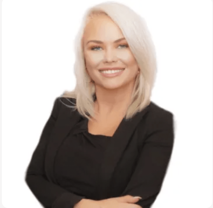 A woman with blonde hair is wearing a black jacket and smiling