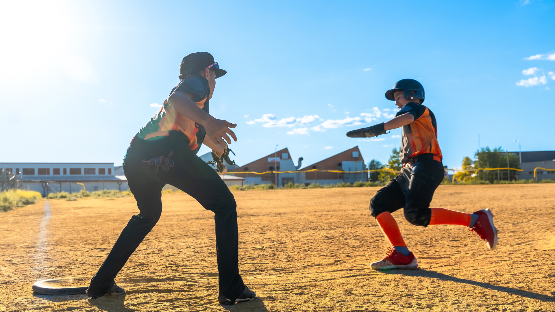 Two softball players in orange jerseys crouch and face each other on a sunny field.
