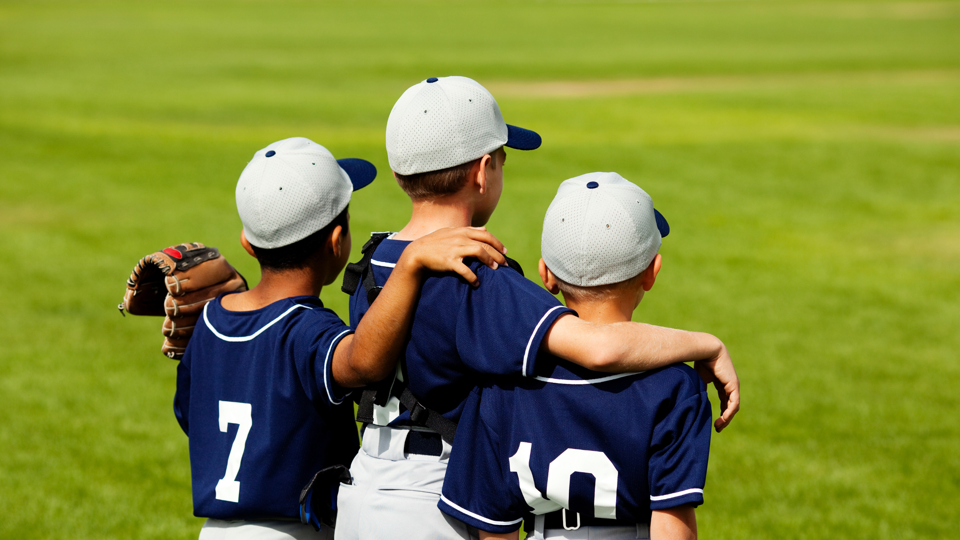 Three youth baseball players in navy jerseys and caps stand arm in arm on a grassy field.