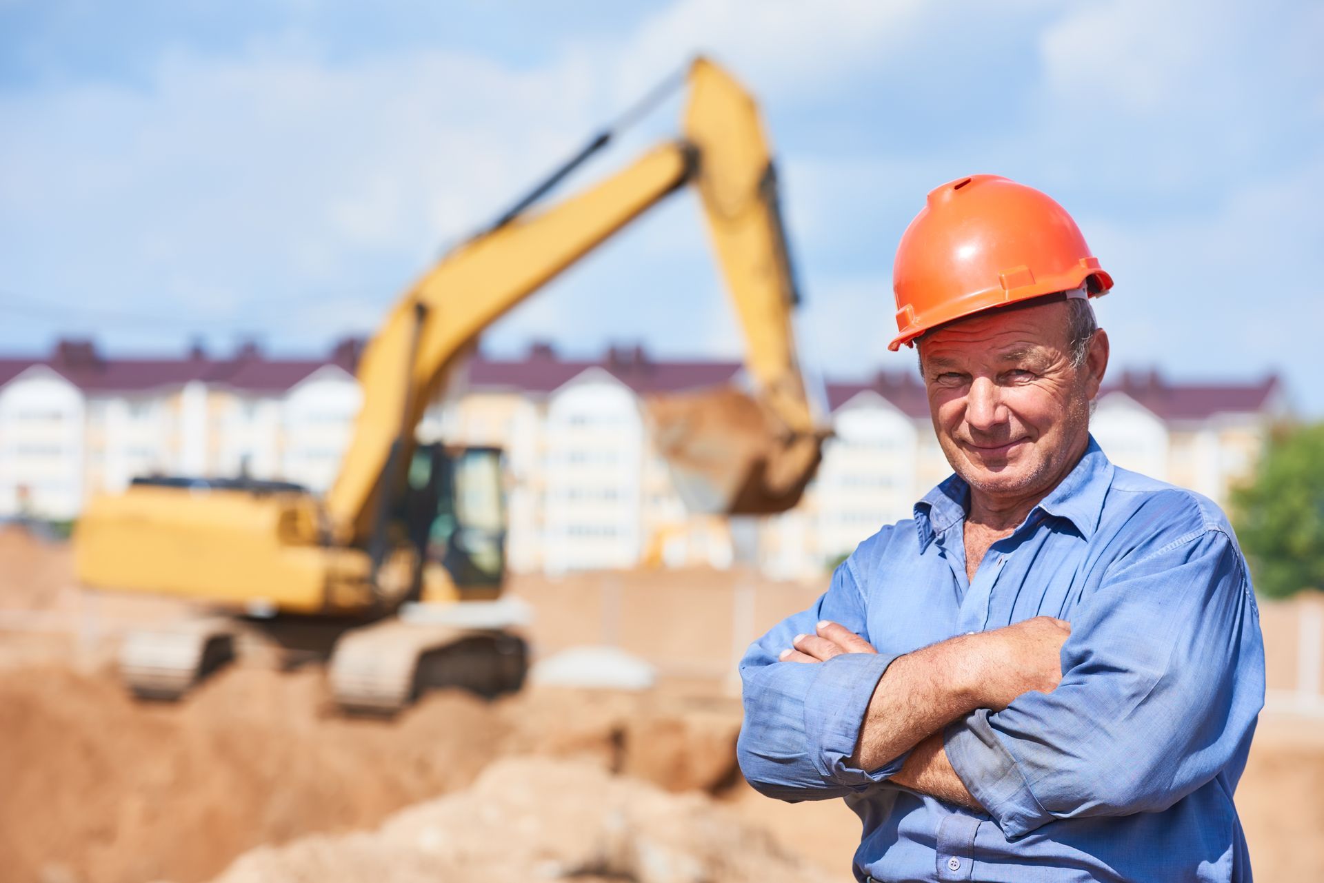 A construction worker looking at the camera with his arms crossed in front of an excavator.
