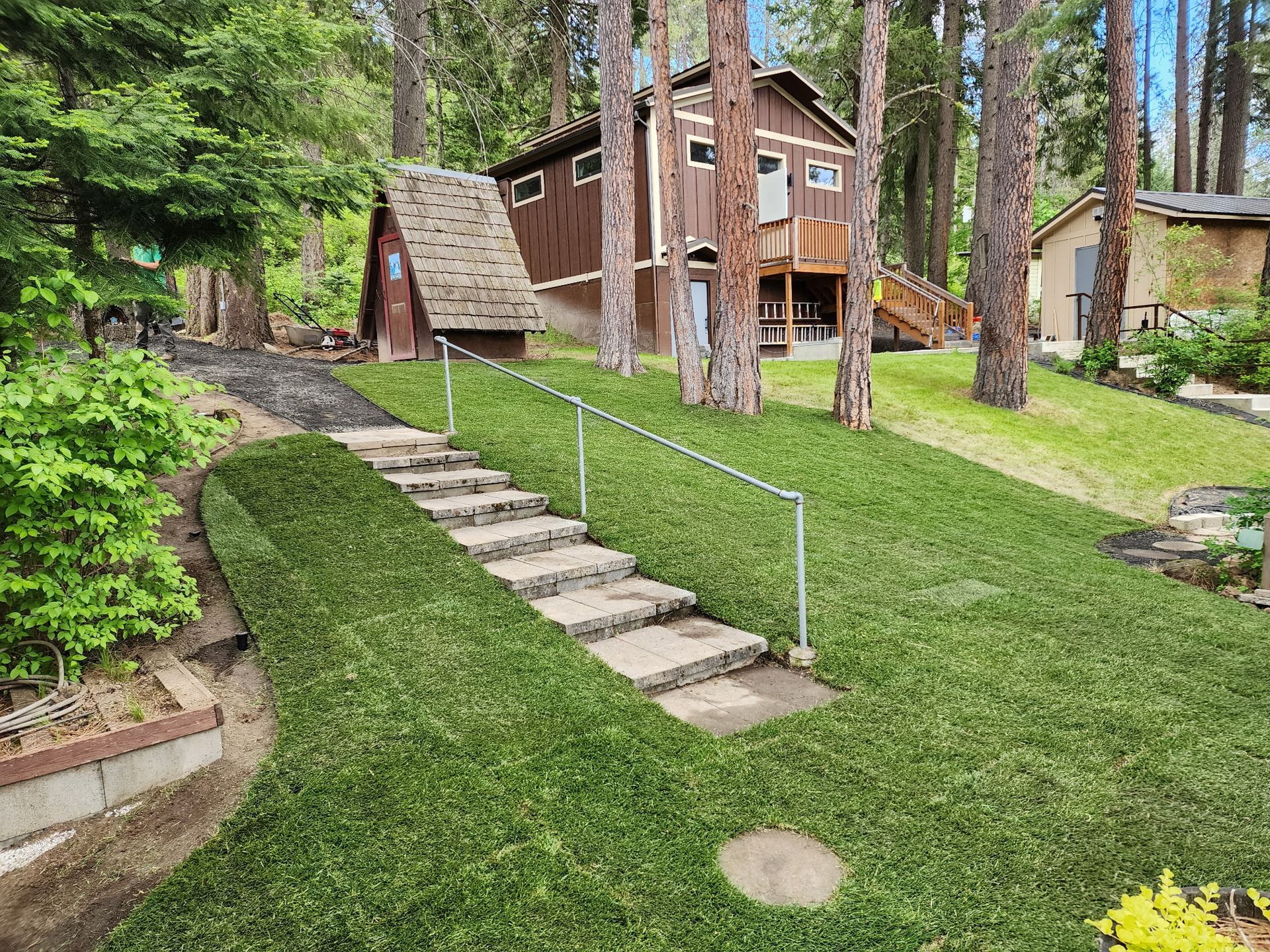 Stone steps lead up to a brown cabin with a wooden deck in a grassy, wooded area.