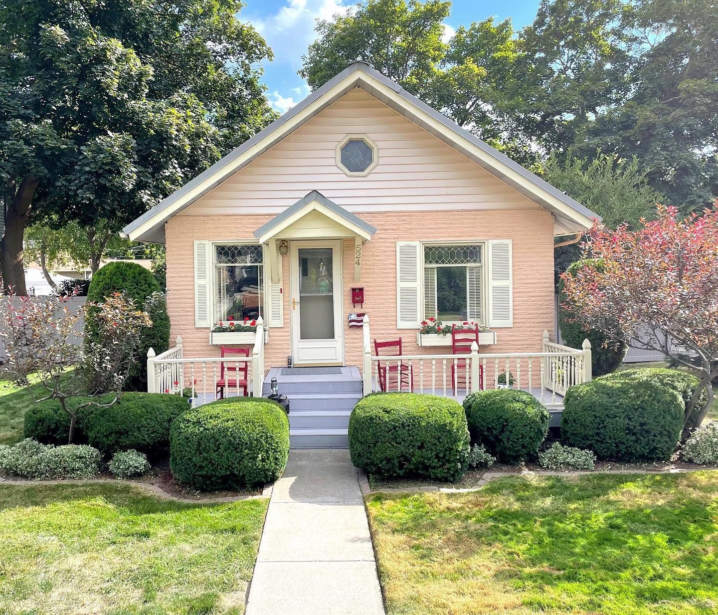 Charming pink brick cottage with white shutters, porch, and red chairs, set in a green yard with manicured bushes.