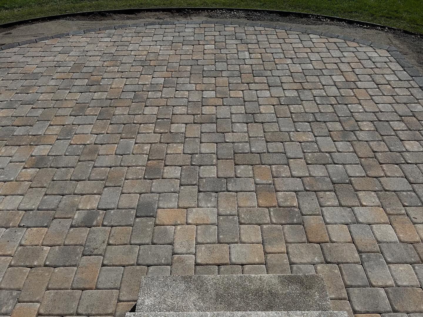 Brick paver patio in varying shades of tan and gray, with a concrete step at the bottom.