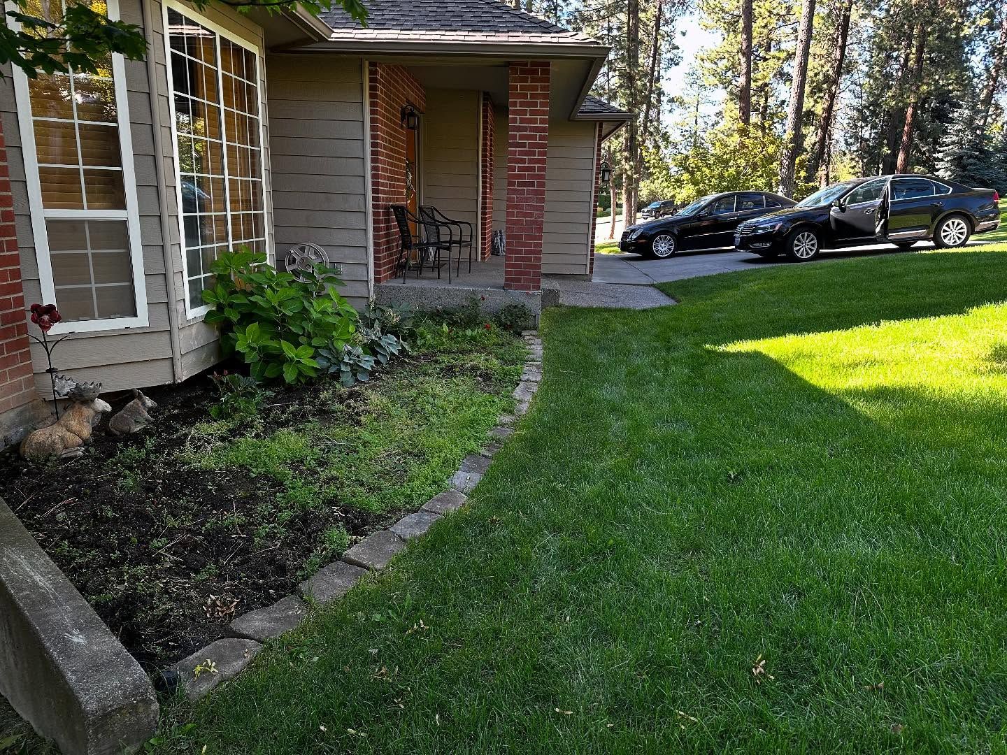 A house with a lawn and garden bed in front. Cars parked on the right, trees in the background.