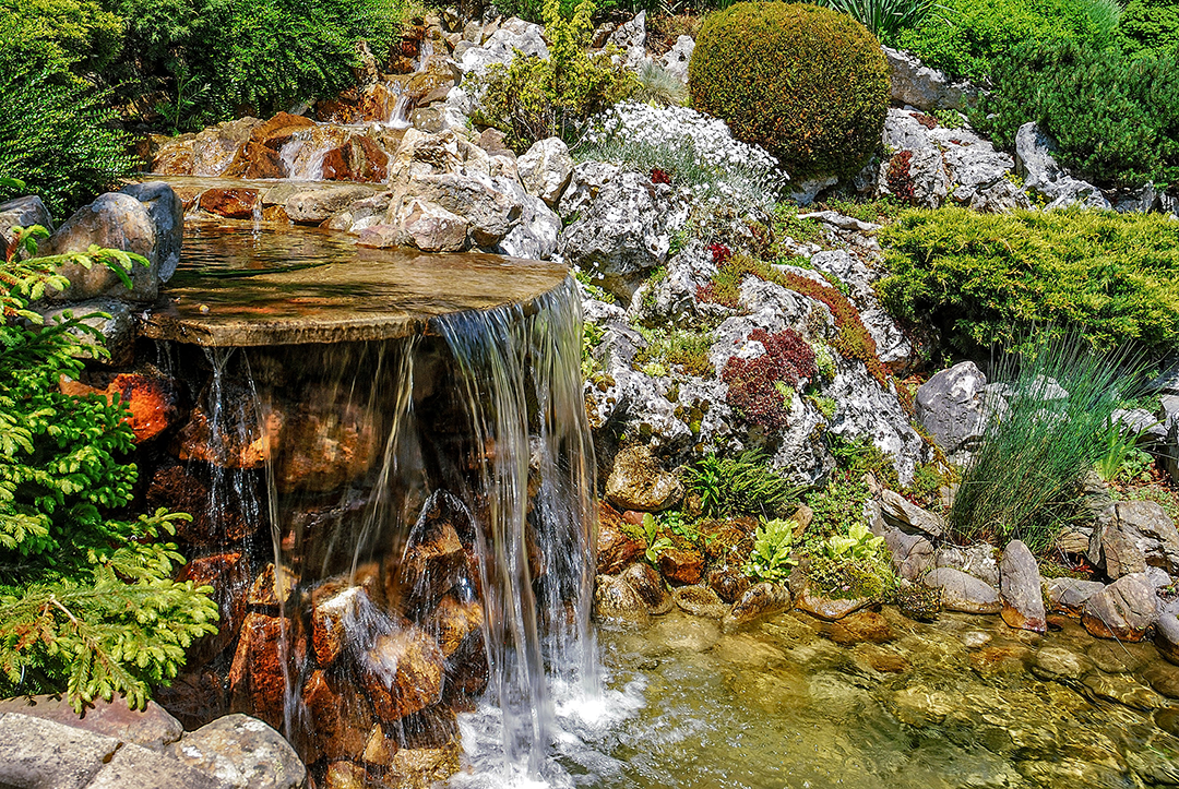 Miniature waterfall cascading over rocks, surrounded by lush greenery and flowers.