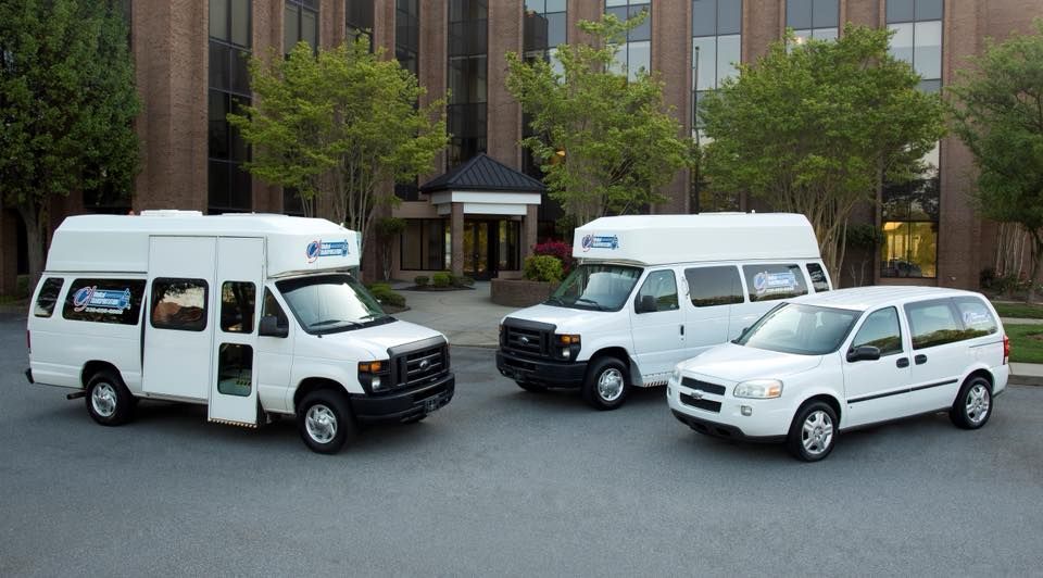 Three white accessible vans parked in front of a building with a tree-lined driveway.