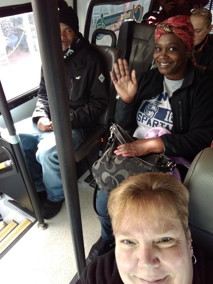 People on a bus: one waving, one with a head wrap, and another taking a selfie.