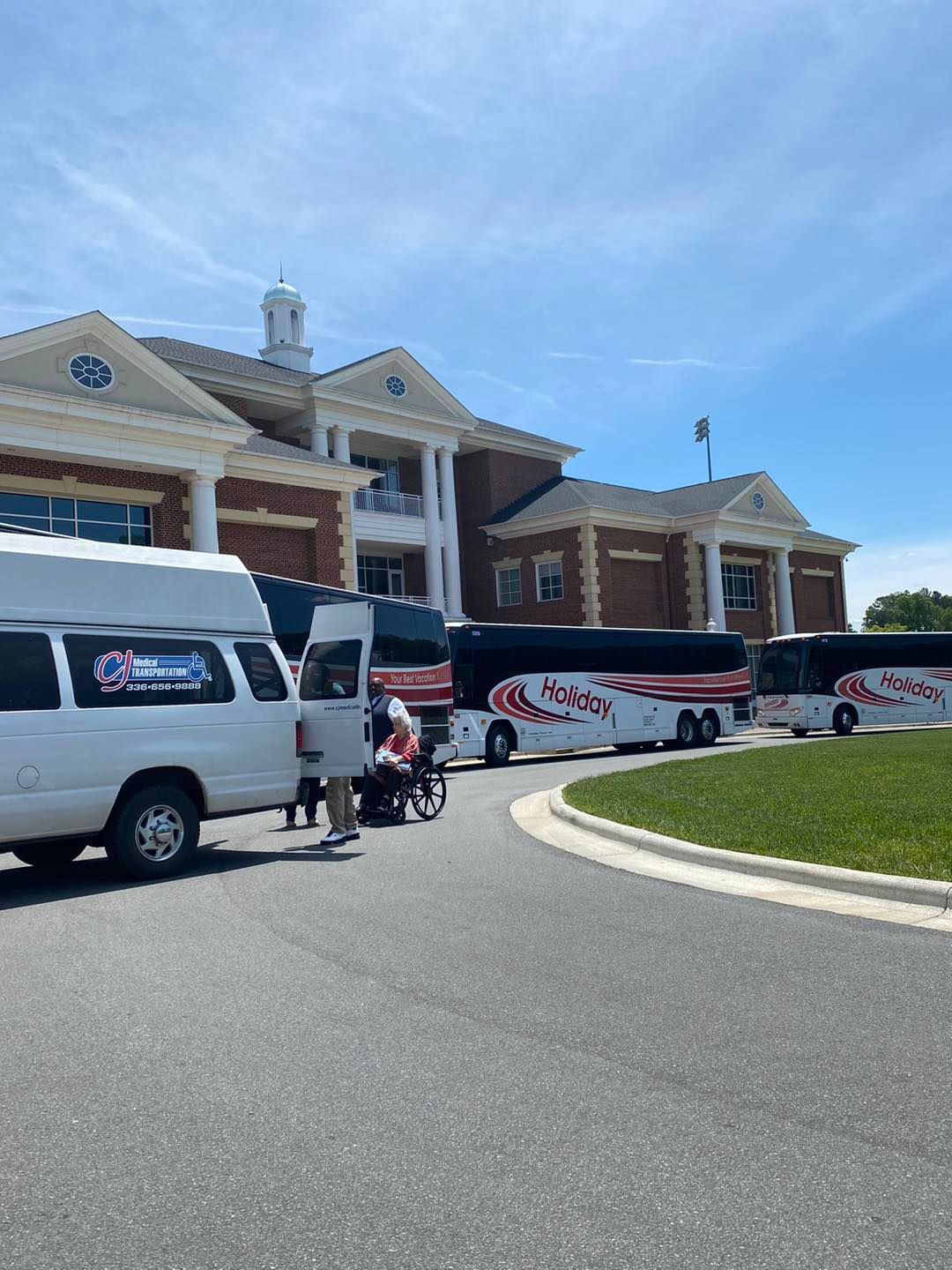 A white accessible van and several tour buses parked near a brick building with columns on a sunny day.