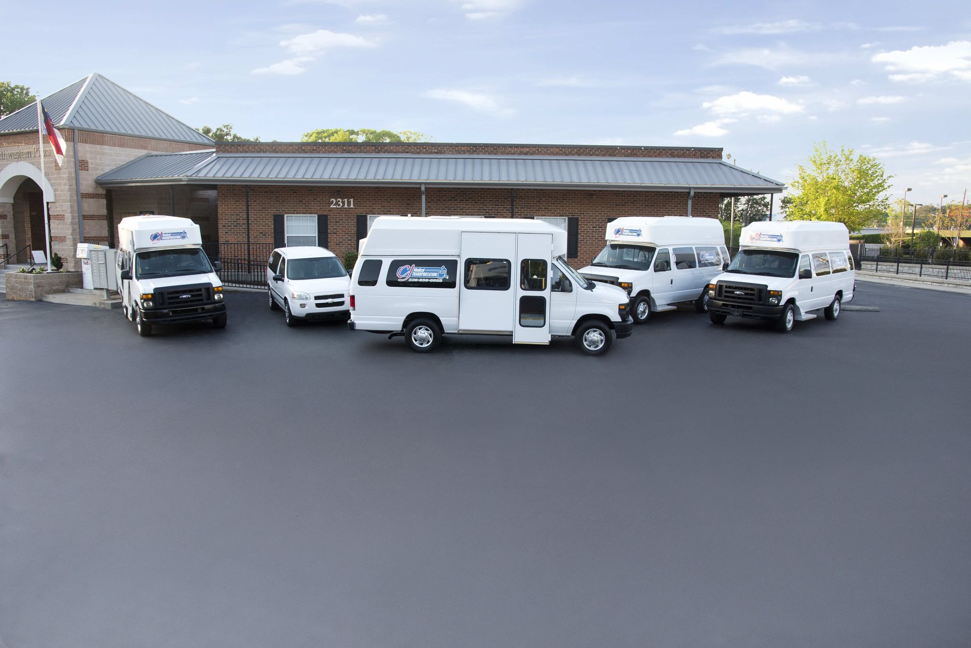 Five white passenger vans parked outside a brick building under a blue sky.