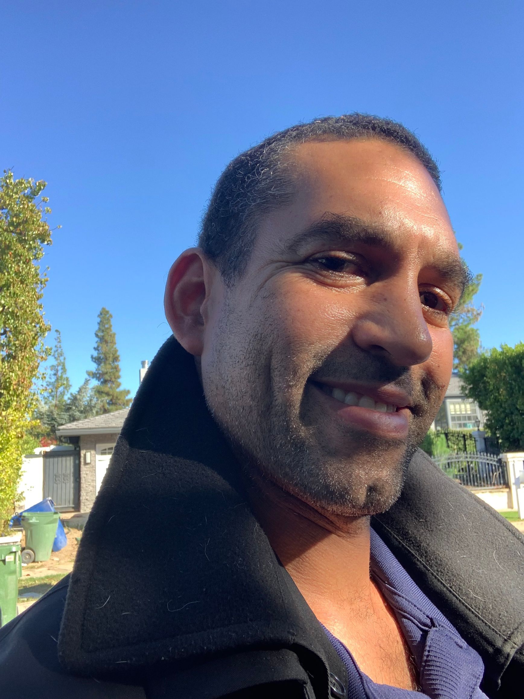 Close-up of a smiling person outdoors in a dark coat with trees and a blue sky in the background