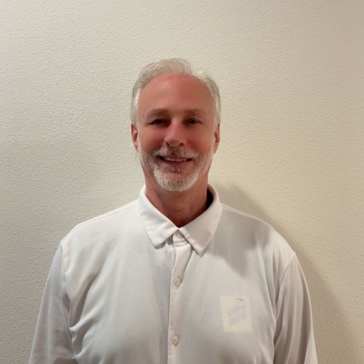 Smiling man in a white button-up shirt standing against a plain beige wall