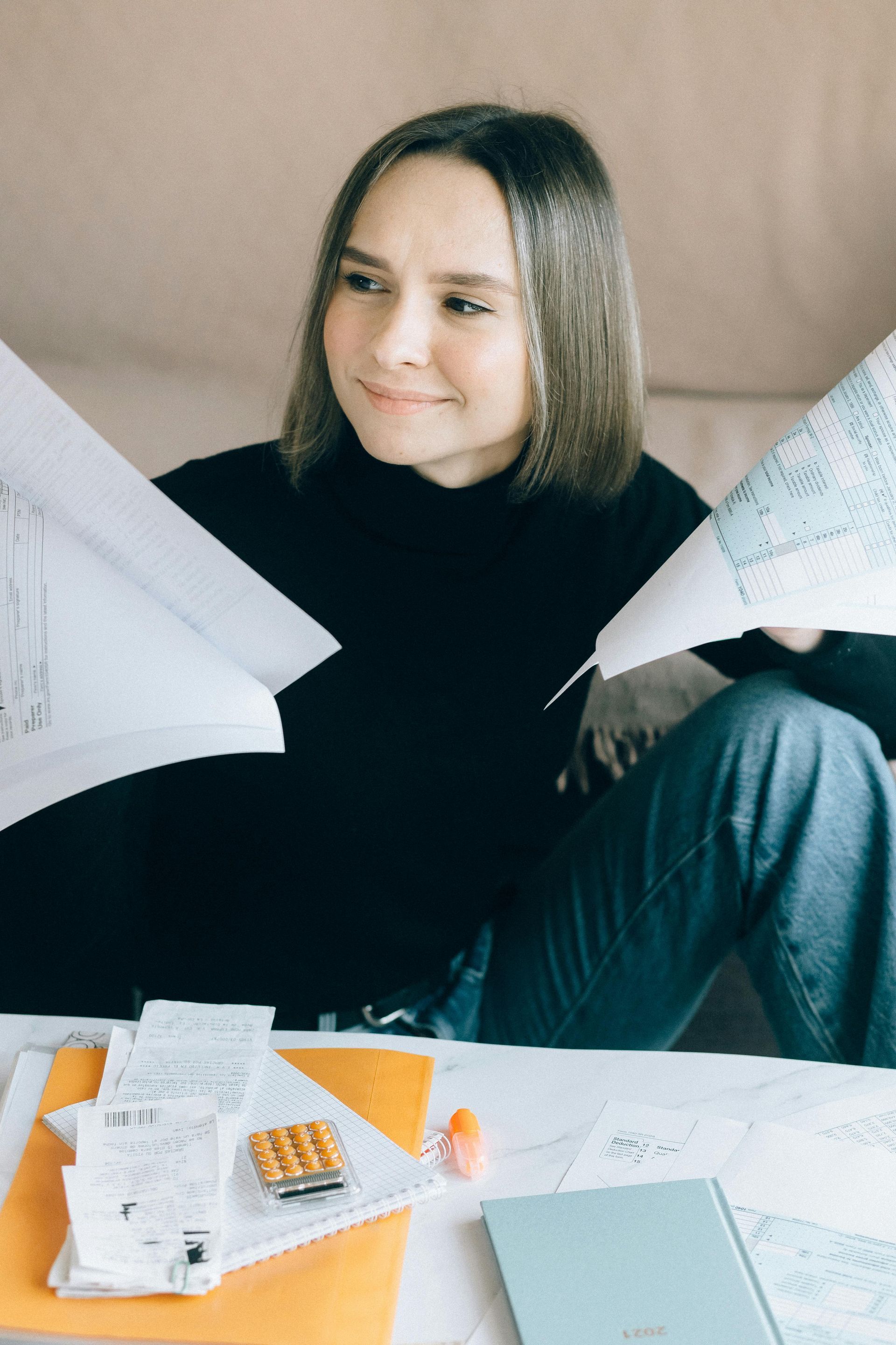 Person seated at a table with papers and calculator, holding two sheets and smiling thoughtfully
