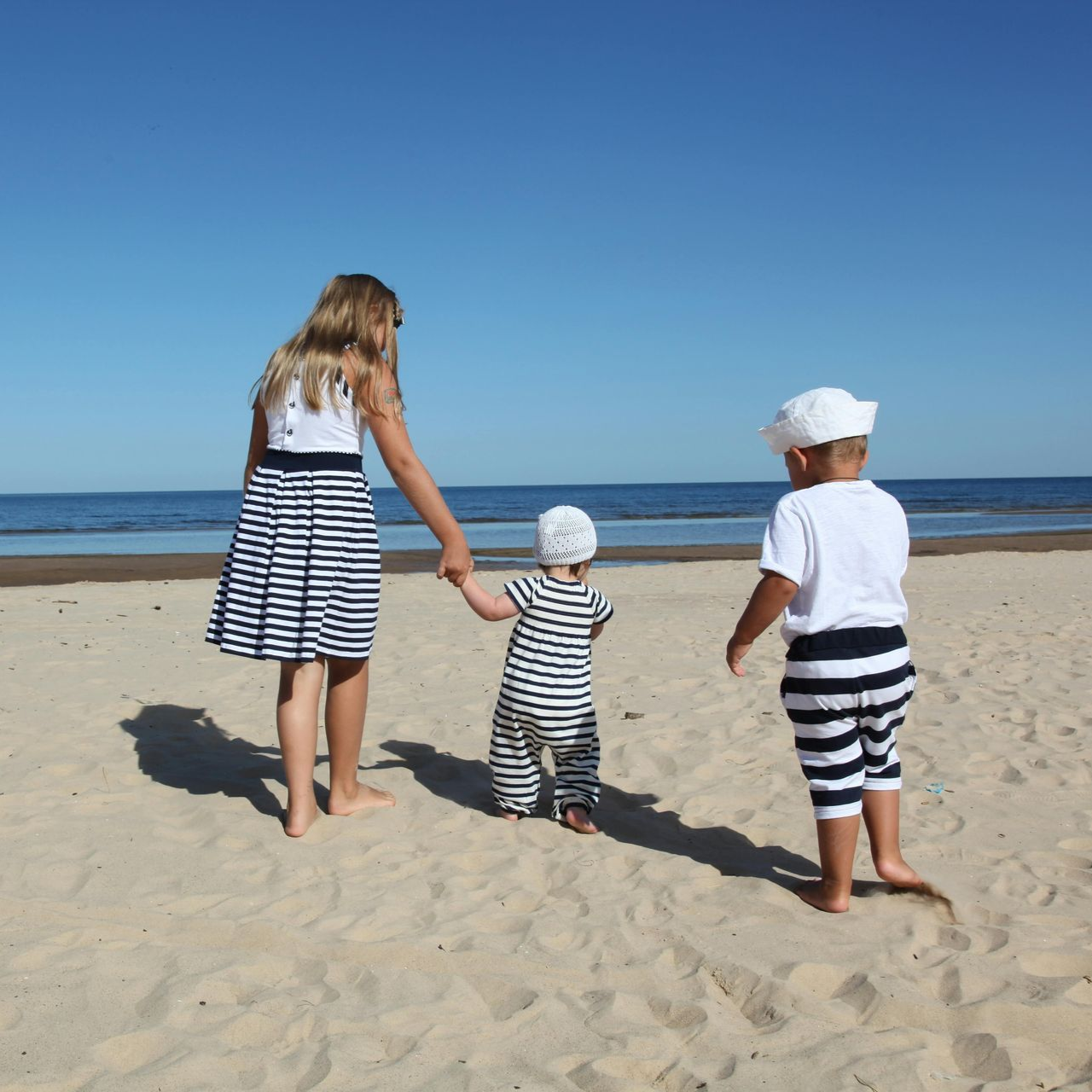 Three children holding hands walking on a sandy beach toward the ocean under a clear blue sky