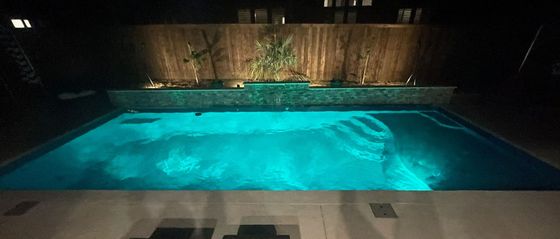 Backyard swimming pool glowing turquoise at night with patio lights and wooden fence behind it