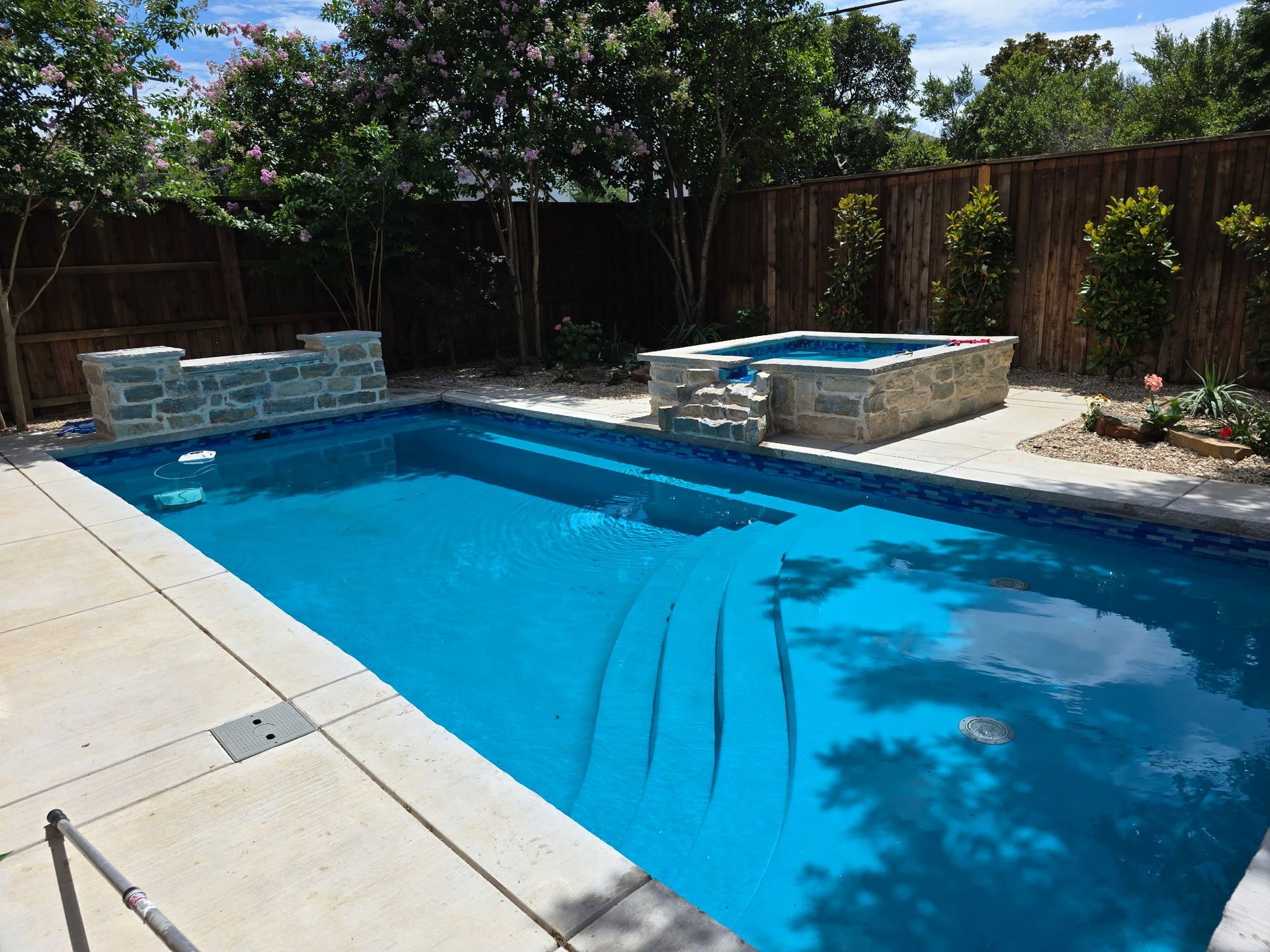 Backyard swimming pool with blue water, stone hot tub, and wooden fence in sunlight