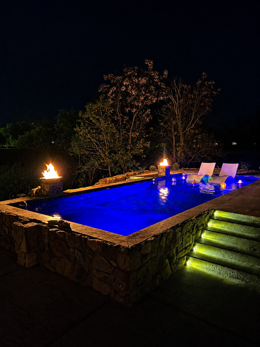 Nighttime pool with blue-lit water, lounge chairs, tiki torches, and glowing steps leading up to it