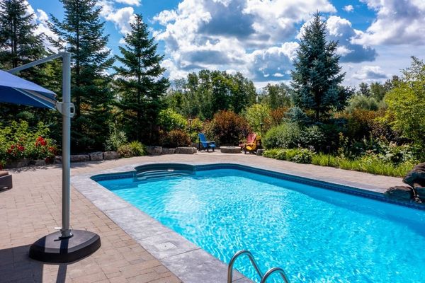 Backyard swimming pool with clear blue water, patio umbrella, and trees under a partly cloudy sky