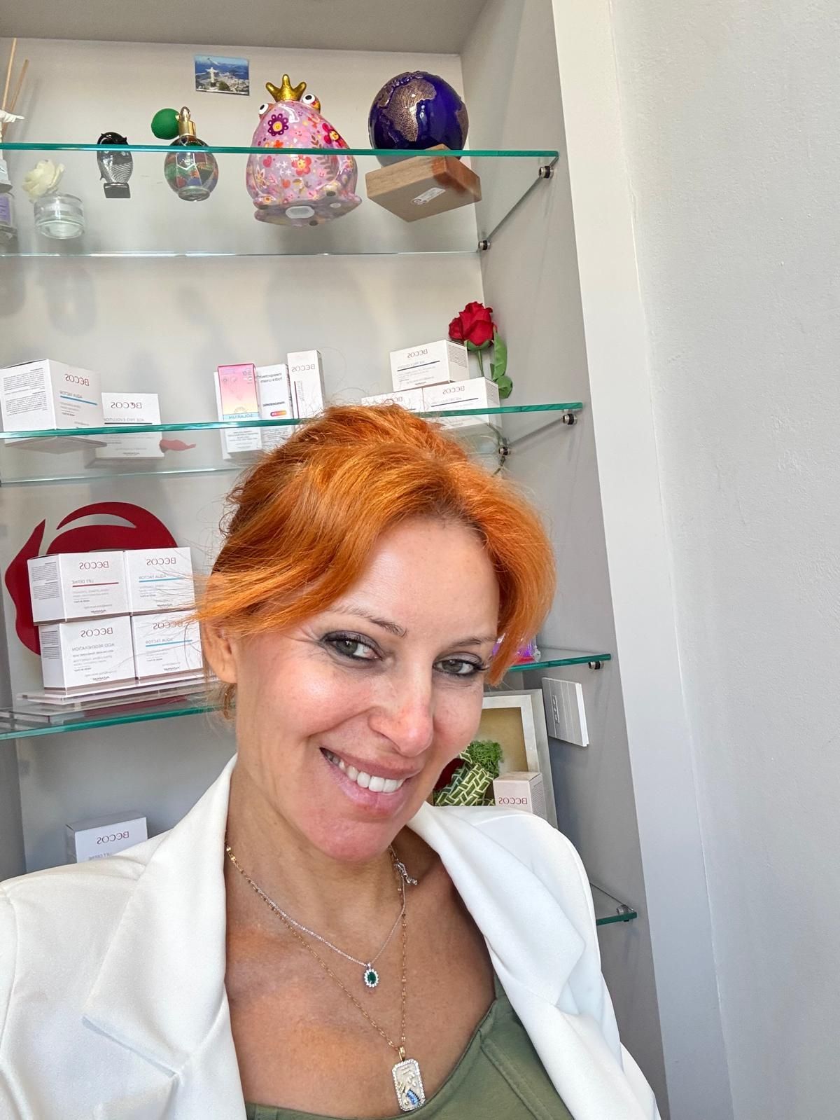 Persona sorridente con i capelli arancioni e una giacca bianca, in piedi davanti a scaffali con oggetti decorativi