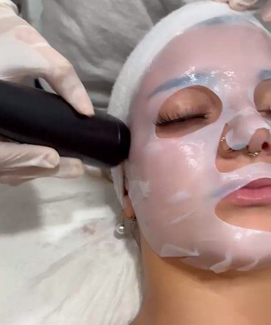 Un trattamento viso con una maschera in tessuto e un dispositivo di bellezza manuale sul viso