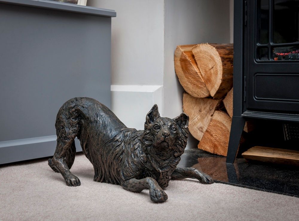 Bronze dog sculpture in playful pose near a stack of logs and a fireplace.
