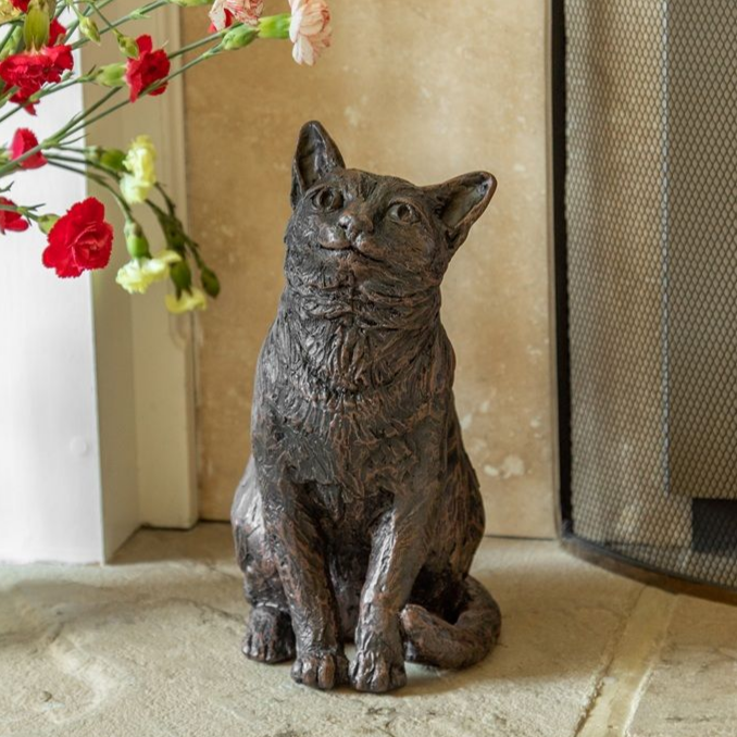 Bronze cat statue seated near flowers and fireplace.