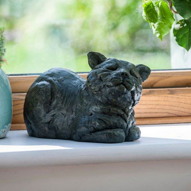 Dark gray cat sculpture resting on a windowsill, with a blurred green background.