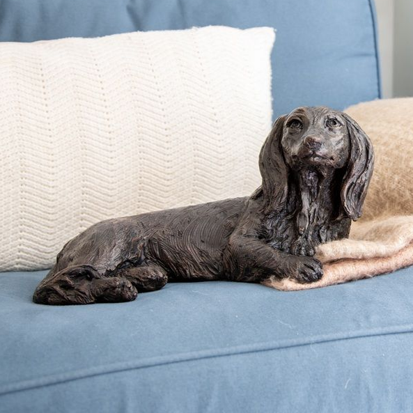 Statue of a dachshund dog lying on a blanket on a blue couch, next to a white pillow.