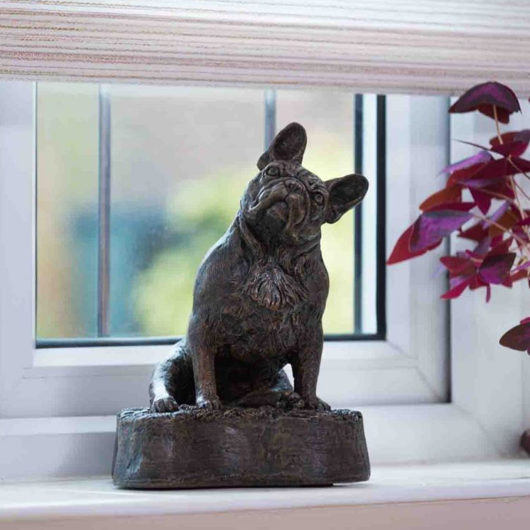 Bronze bulldog statue on a windowsill, looking upwards with a curious expression, near a window with foliage.