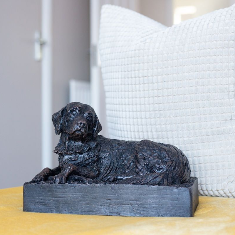 Black dog sculpture, reclining on a rectangular base, next to a textured white pillow.