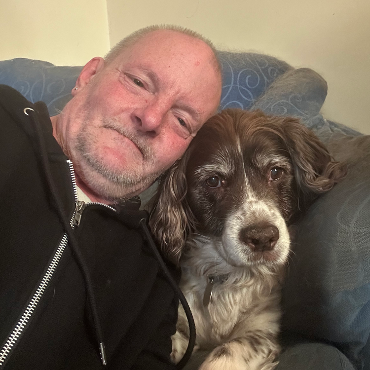 Man in black jacket cuddles with brown and white dog on a blue couch.