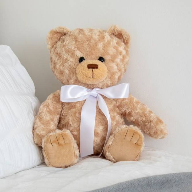 Tan teddy bear with a white bow tie, sitting on a bed next to a white pillow.