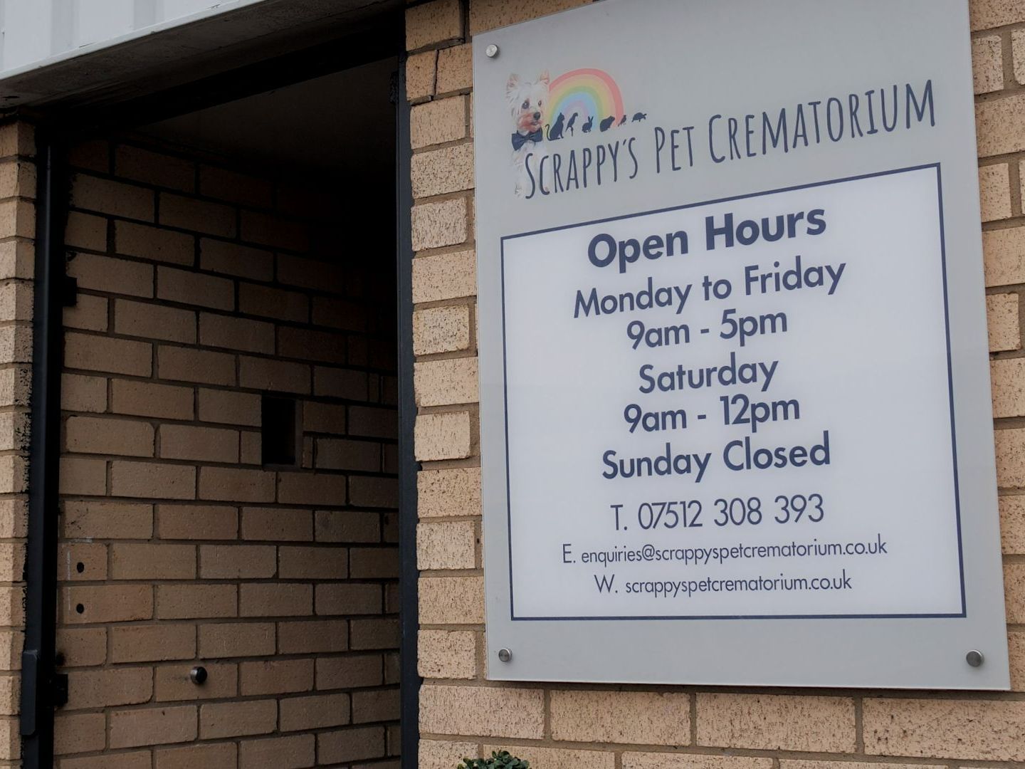 Sign for Scrappy's Pet Crematorium displaying open hours and contact information on a brick building.