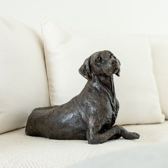 Bronze dog sculpture on a white couch. The dog is lying down, looking up.