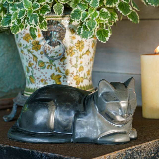 Silver cat figurine resting on a wooden surface, with a floral pot and candle in the background.