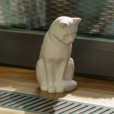 White ceramic cat statue, sitting, looking down. Sitting on wooden ledge, near window and vent.