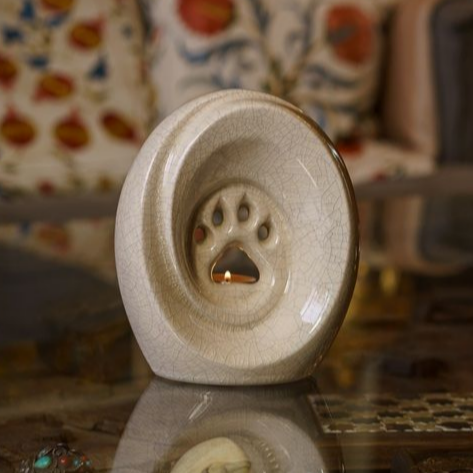 Cream-colored pet cremation urn with a paw print cutout, glowing candle inside, on reflective surface.
