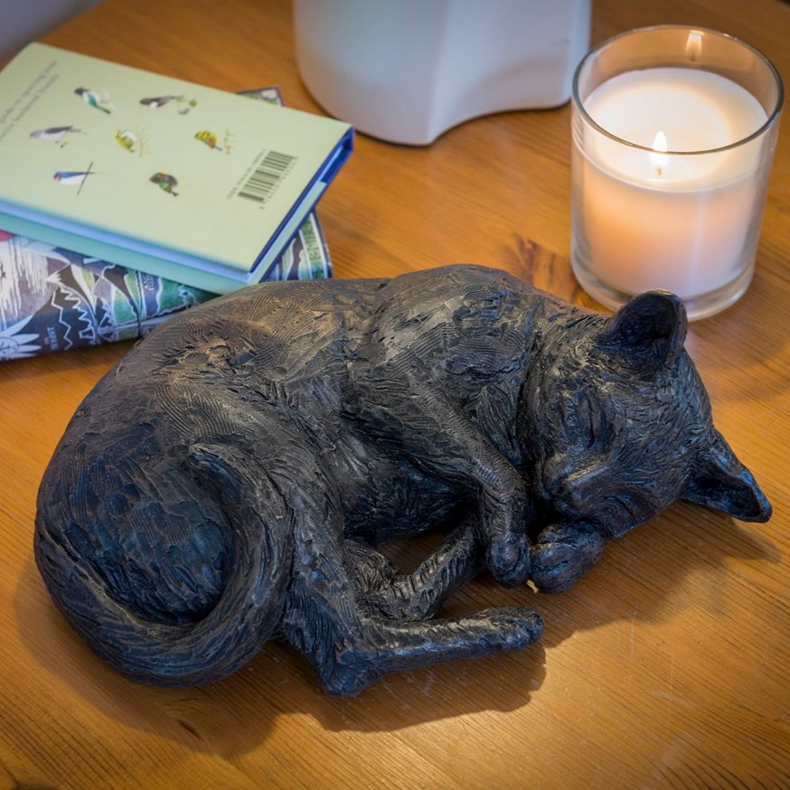 Sleeping black cat sculpture on a wooden table, next to books and a lit candle.