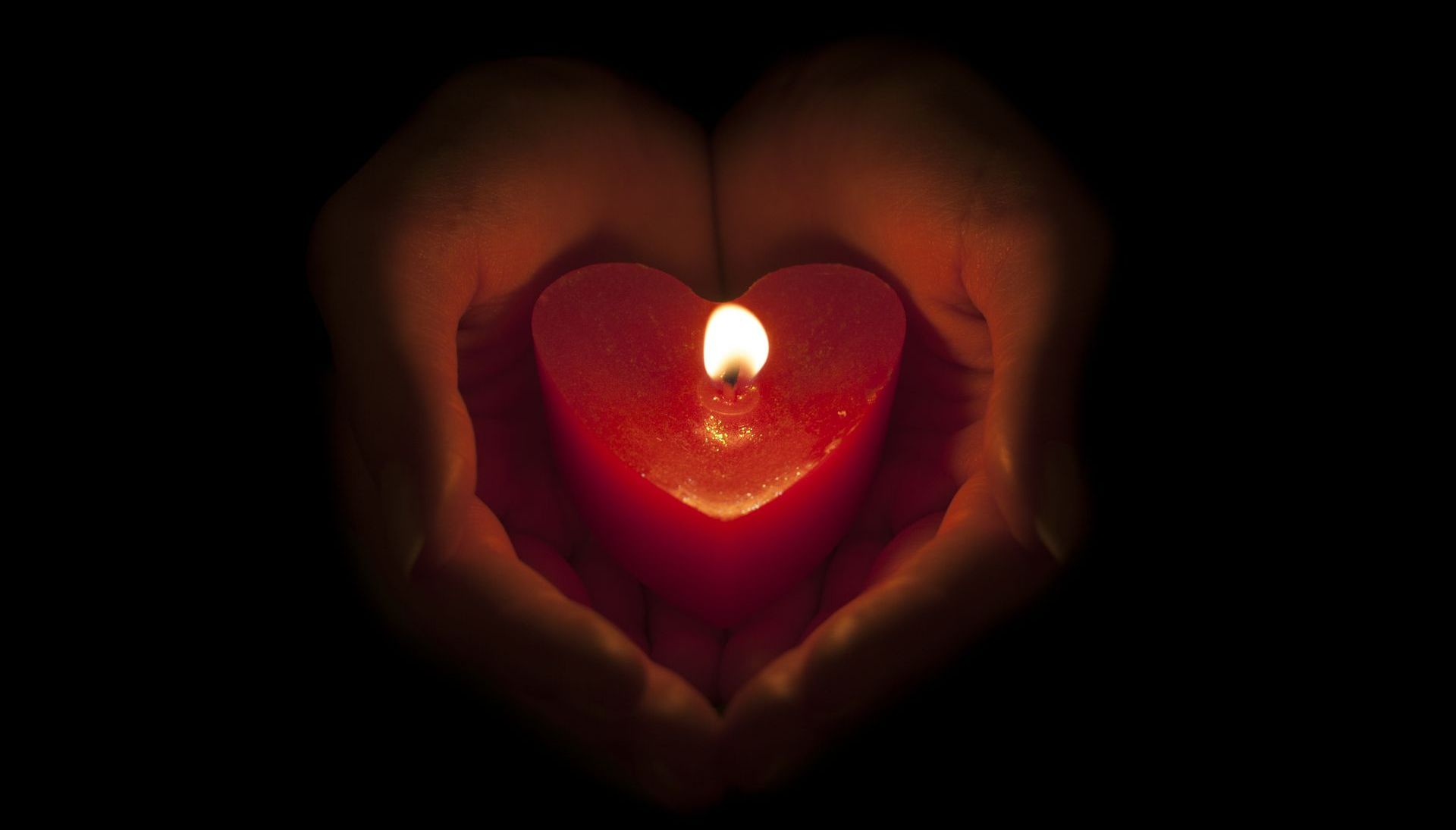 Hands cupping a lit, heart-shaped red candle against a dark background.