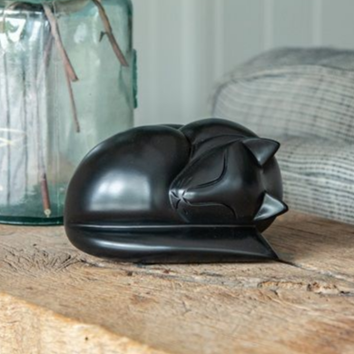 Black cat sculpture curled up on a wooden surface, with a glass vase and a patterned pillow in the background.