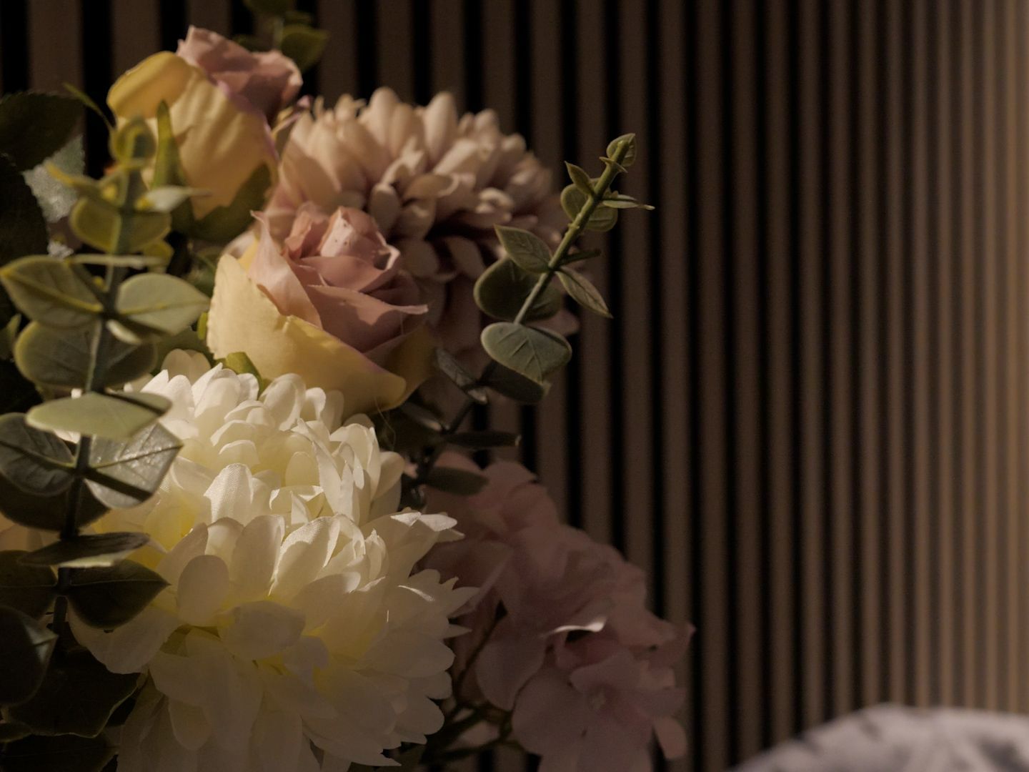 Close-up of a bouquet with white, pink, and yellow flowers in front of a striped brown and beige wall.