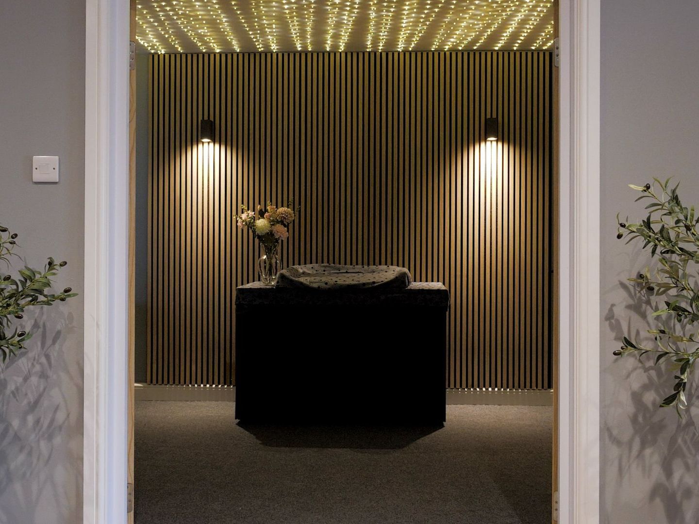 Dark room interior with table, floral arrangement, and vertical wood panel backdrop lit by wall sconces.