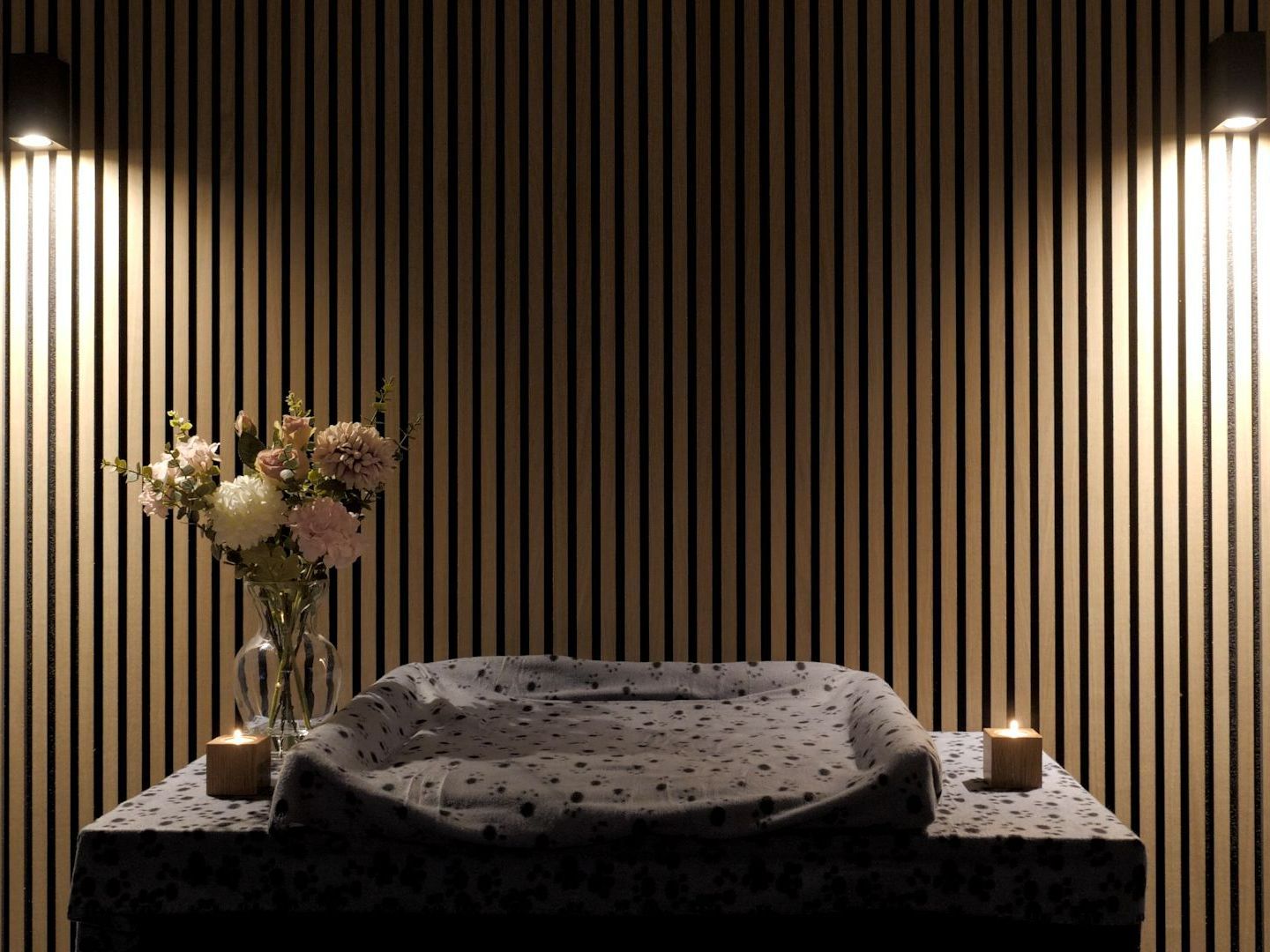 Wooden-slat wall with a table holding flowers, a baby changing pad, and candles, lit by spotlights.