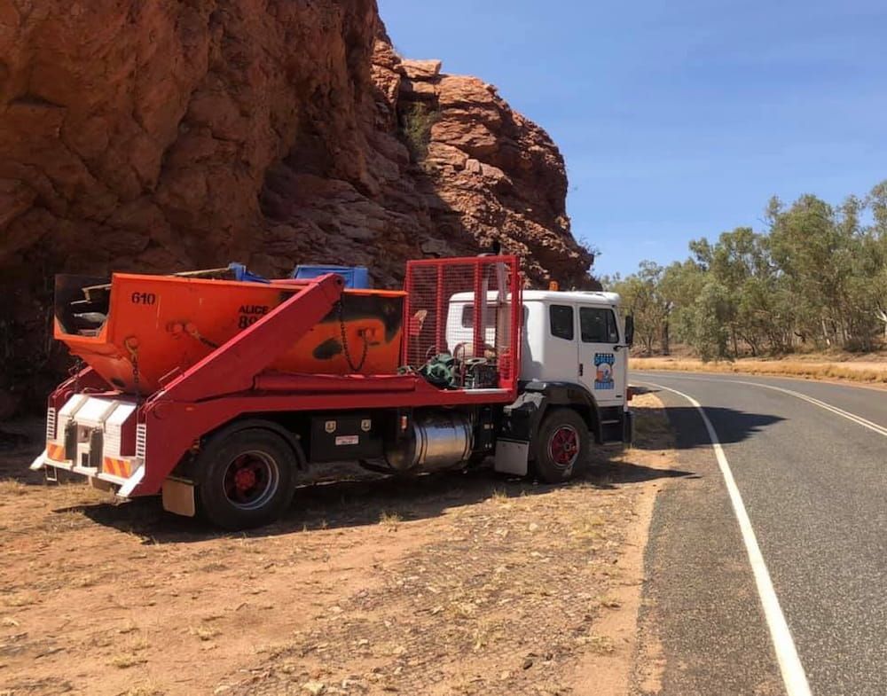 A white truck with an orange skip bin parked on the dirt shoulder of a road next to a large, reddish-brown rock cliff. — Alice Skip Hire in Alice Springs, NT