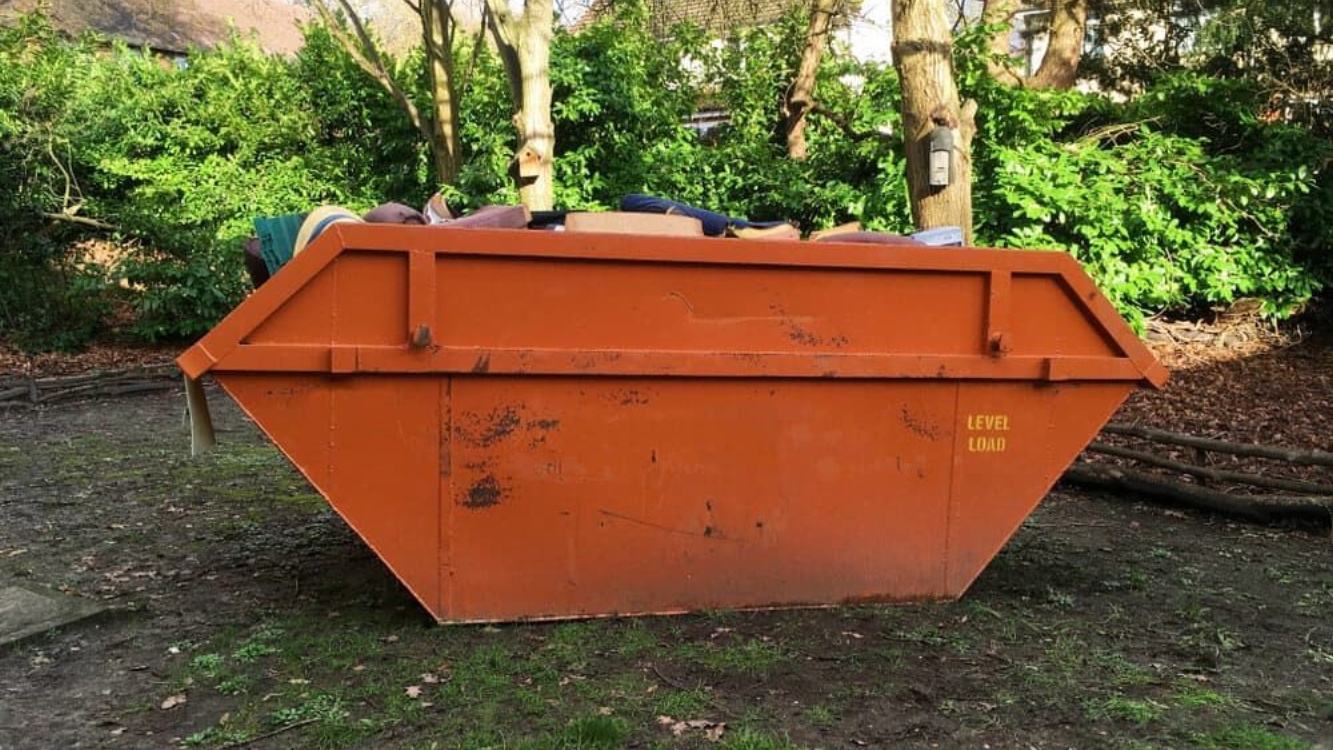 A bright orange, rectangular construction skip filled with mixed debris, situated on a grassy area in front of trees. — Alice Skip Hire in Alice Springs, NT 