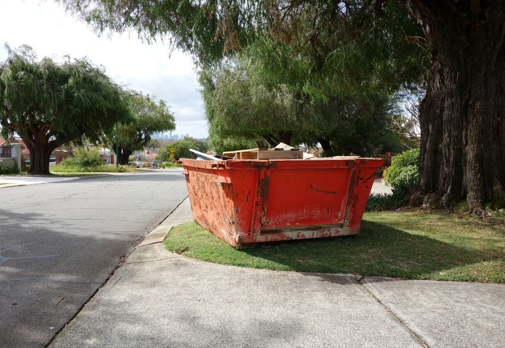 Large orange skip bin on a grassy curb beside a sidewalk and road under trees — Alice Skip Hire in Alice Springs, NT