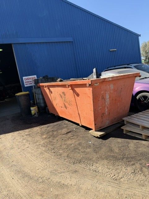 Large orange dumpster beside a blue metal building on a dirt lot, with a car partially visible nearby. — Alice Skip Hire in Alice Springs, NT