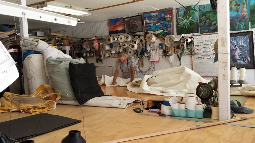 A person works on a large piece of white fabric in a workshop filled with sewing supplies and wall-mounted art.