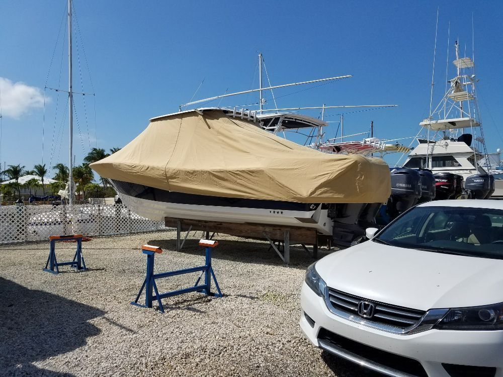 A boat with a tan cover sits on jack stands in a gravel lot next to a white Honda car on a sunny day.