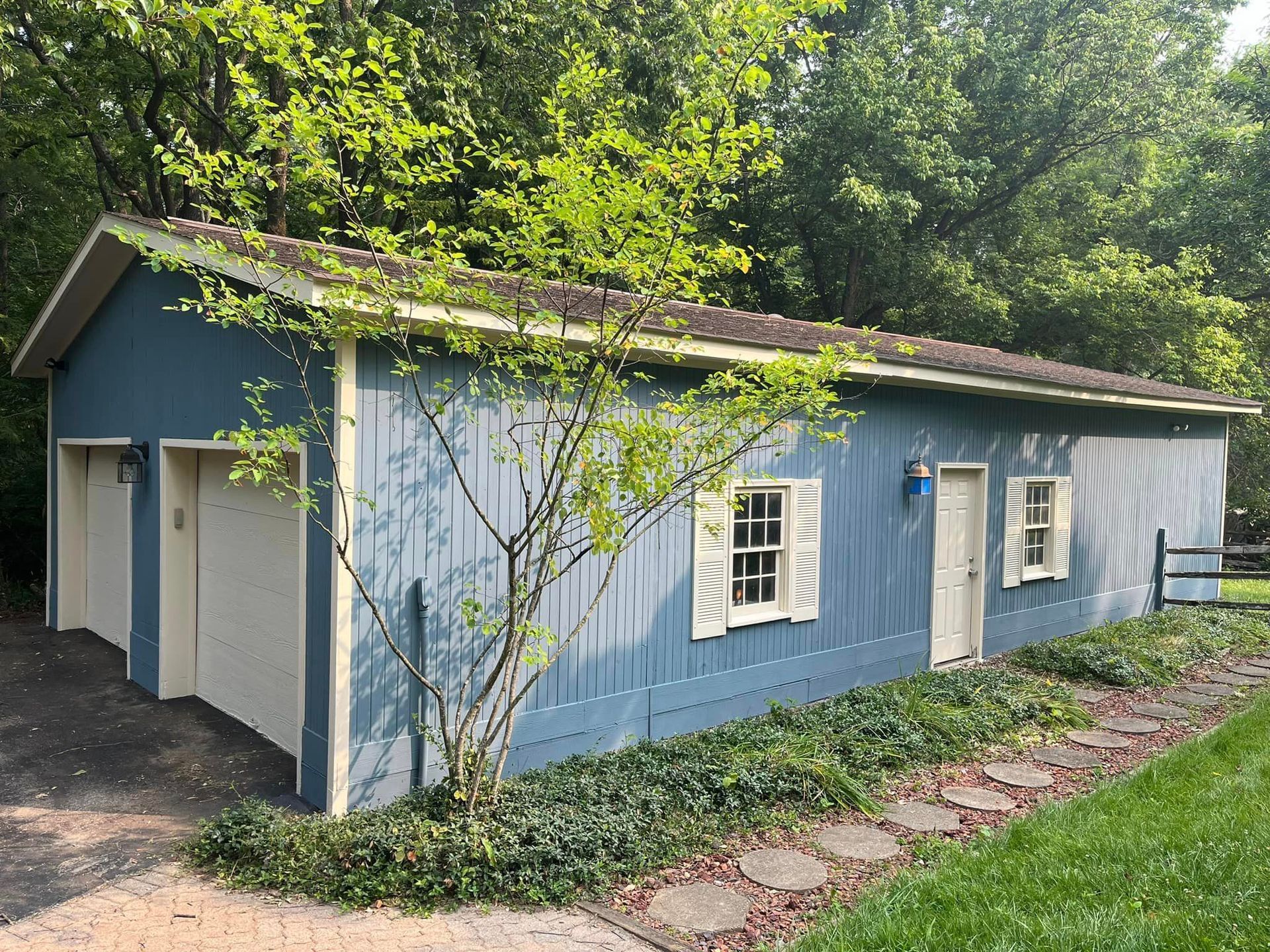 A blue garage with white shutters is surrounded by trees and grass.