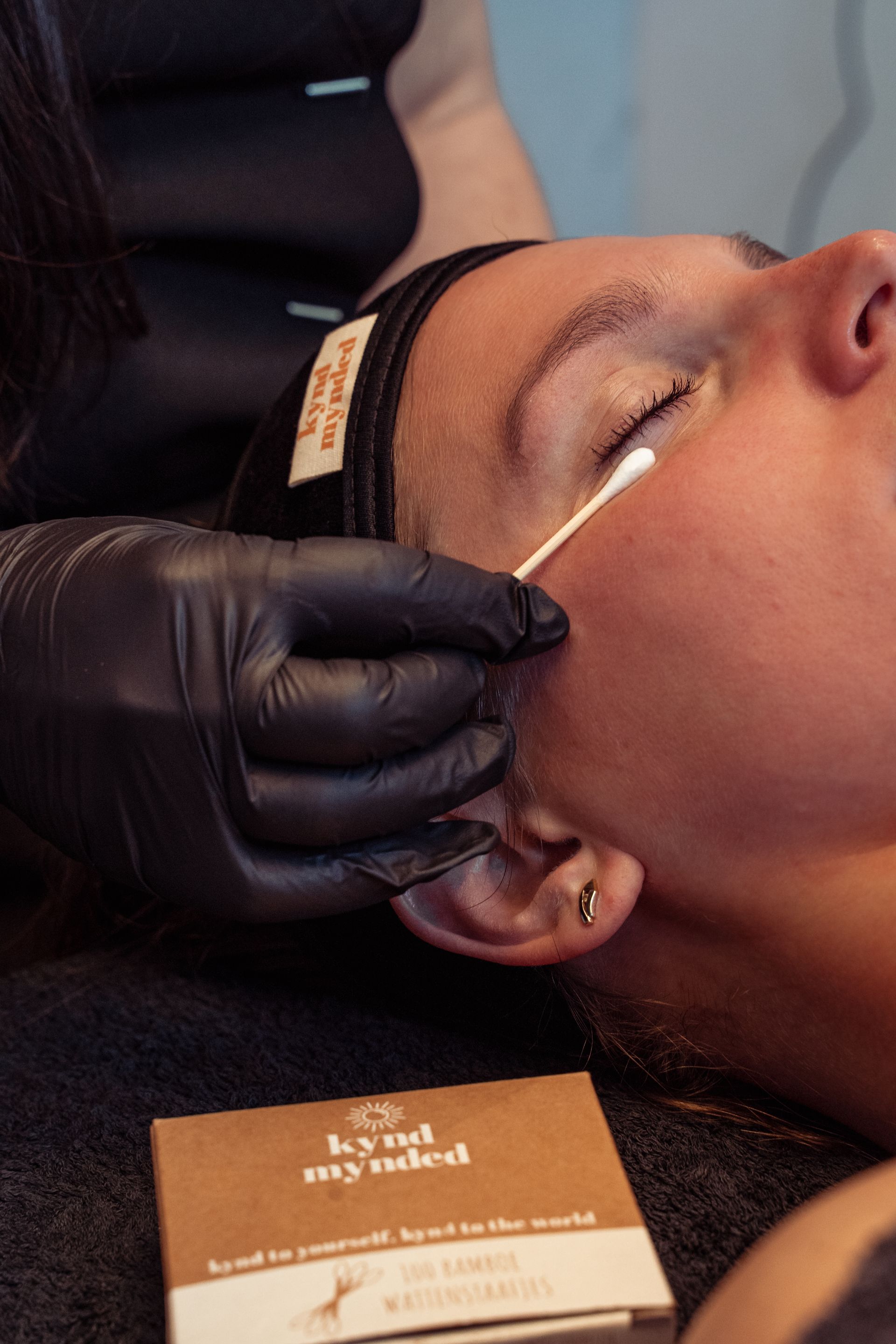 Tijdens een gezichtsbehandeling in een spa brengt een gehandschoende hand met een wattenstaafje een behandeling aan op de huid rond de gesloten ogen van de patiënt.